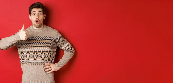 Image of handsome caucasian man in christmas sweater looking amazed, showing thumb-up in approval, like something, standing over red background.