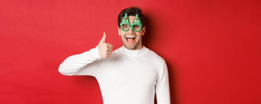 Concept of christmas, winter holidays and celebration. Excited handsome guy in party glasses, smiling amused and showing thumb-up, standing over red background.