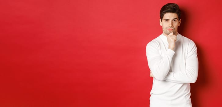 Image of thoughtful handsome man making assumption, thinking and looking at camera, standing in white sweater against red background.