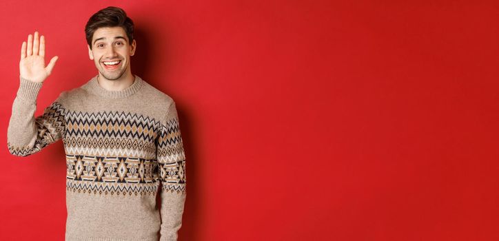 Portrait of handsome and friendly guy in christmas sweater, saying hello and waving hand, greeting someone and wishing happy winter holidays, standing over red background.