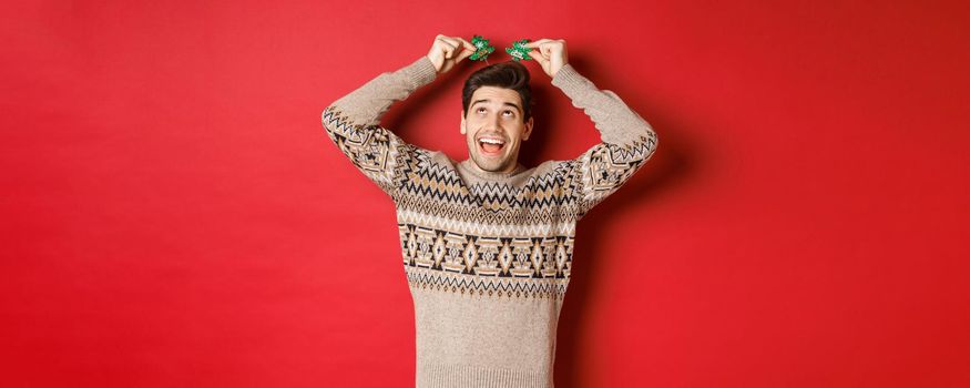 Concept of winter holidays, christmas and celebration. Image of funny and cute adult man enjoying new year party, smiling and rejoicing, standing over red background.