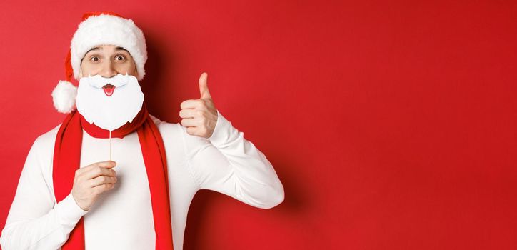 Concept of christmas, winter holidays and celebration. Portrait of funny man in santa hat, holding beard mask, showing thumb-up, enjoying new year party, standing over red background.