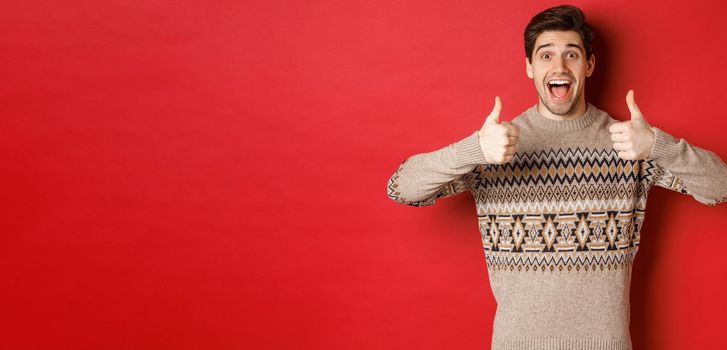 Portrait of cheerful man in christmas sweater like something good, showing thumbs-up and smiling amazed, praise new year party, standing over red background.