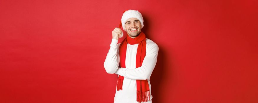Portrait of happy man enjoying christmas holidays, wearing santa hat and scarf, smiling joyfully, standing over red background.