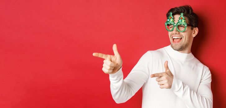 Close-up of cheeky handsome man in party glasses, pointing finger left and greeting person, wishing merry christmas, standing over red background.
