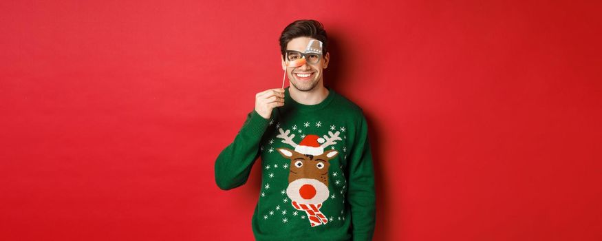 Funny man in christmas sweater and party mask, celebrating winter holidays, smiling happy, standing over red background.