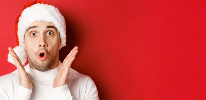 Close-up of amazed handsome man in santa hat and white sweater, raising hands and looking impressed, standing over red background.