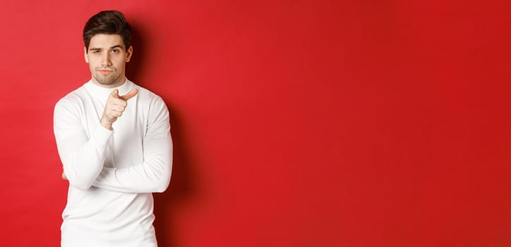 Portrait of thoughtful handsome man in white sweater, pointing at camera and making choice, standing against red background.