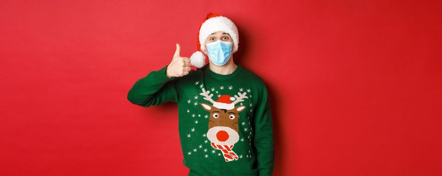 Concept of new year, covid-19 and social distancing. Amazed young guy celebrating christmas, wearing medical mask and santa hat, showing thumb-up in approval, red background.
