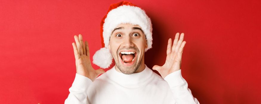 Close-up of happy young man in santa hat, making big christmas announcement, smiling amazed, standing over red background.