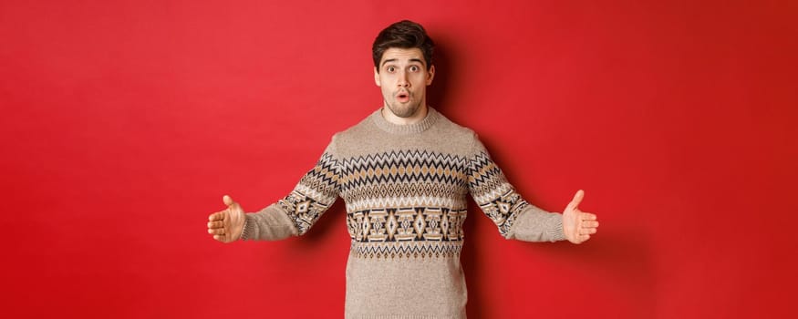 Image of amazed handsome man showing size of christmas present, spread hands sideways and shaping something large, standing over red background.