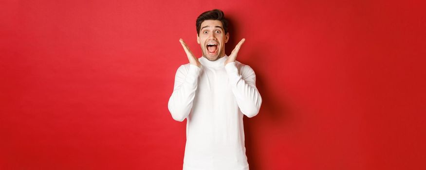 Concept of winter holidays, christmas and lifestyle. Surprised handsome man in white sweater, reacting to amazing new year promo offer, screaming amazed and raising hands, red background.