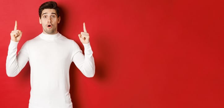 Concept of winter holidays. Good-looking young man with bristle, wearing white sweater, showing christmas advertisement on copy space, pointing fingers up and looking surprised, red background.