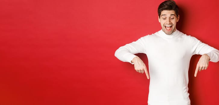 Portrait of happy good-looking man, pointing fingers down and looking at advertisement with excited smile, standing over red background.