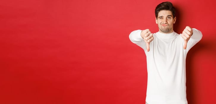 Portrait of disappointed skeptical guy, wearing white sweater, showing thumbs-down to disagree, express dislike, standing over red background.