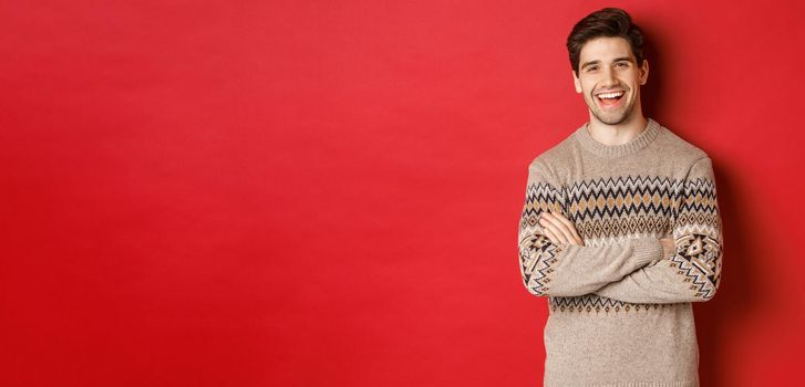 Portrait of cheerful, attractive man in christmas sweater, laughing and smiling, celebrating new year and winter holidays, standing over red background.