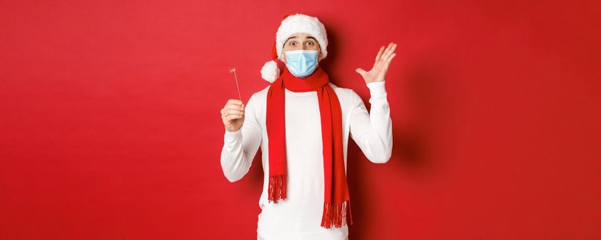 Concept of covid-19, christmas and holidays during pandemic. Cheerful handsome man in medical mask and santa hat, celebrating new year with sparkler, looking excited, red background.