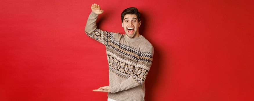 Portrait of excited and happy handsome man in christmas sweater, showing something big, holding large gift for holidays, standing over red background.