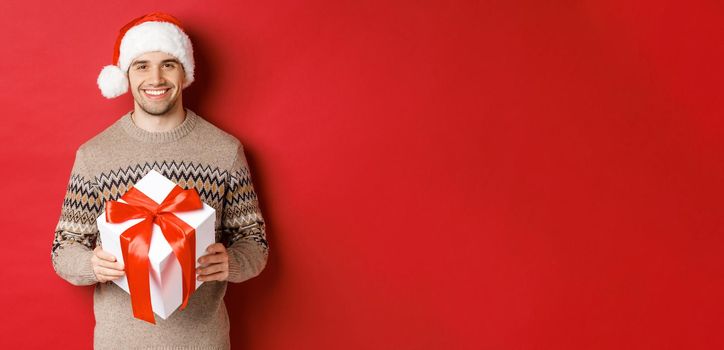 Image of handsome bearded man in winter sweater and santa hat, holding christmas gift and smiling, have a present wrapped with ribbon, standing over red background.