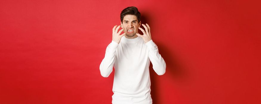 Image of angry and pissed-off man in white sweater, grimacing and shaking from rage, standing furious against red background.