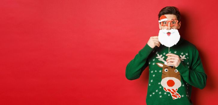 Portrait of surprised man in green christmas sweater, holding funny santa claus mask, celebrating new year, standing over red background.