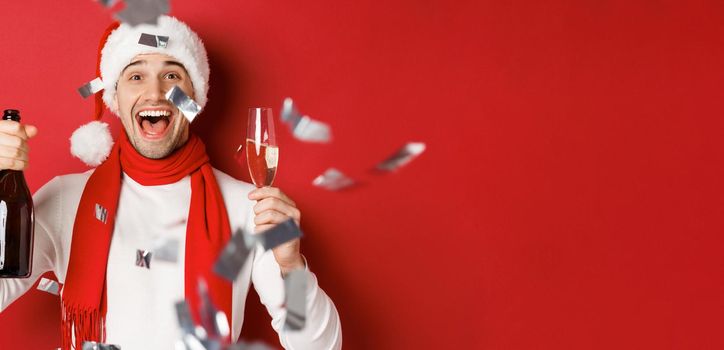 Concept of winter holidays, christmas and lifestyle. Close-up of happy man celebrating new year, holding champagne bottle and glass, standing over red background with confetti.