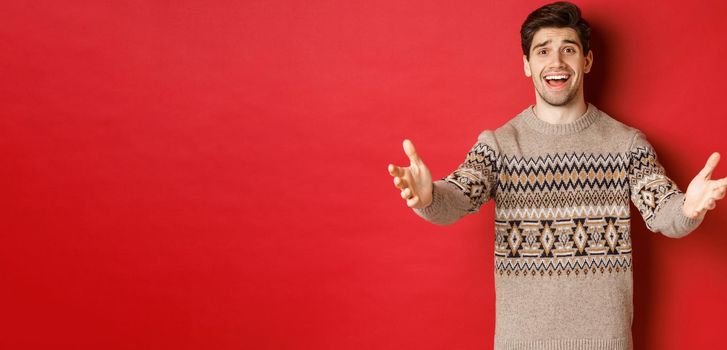 Image of happy and flattered handsome guy in christmas sweater, reaching hands to welcome guests on new year party, inviting to celebration, standing over red background.