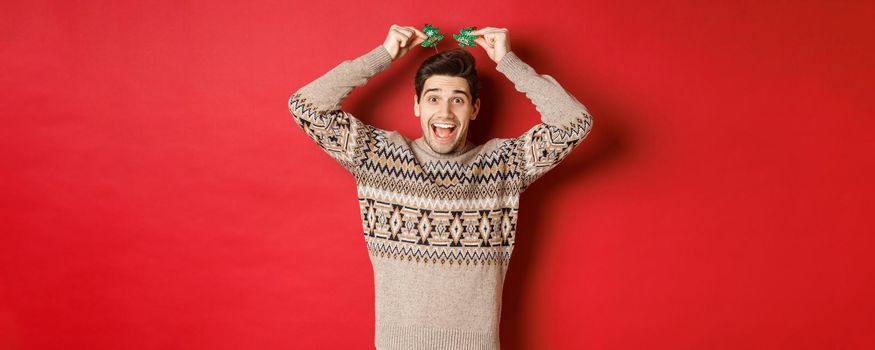 Concept of winter holidays, christmas and celebration. Image of funny and cute adult man enjoying new year party, smiling and rejoicing, standing over red background.