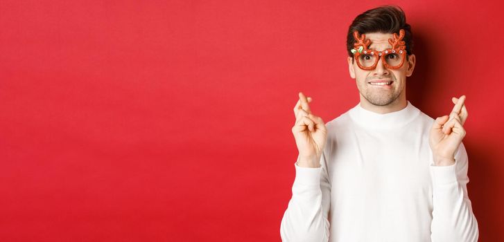 Close-up of handsome nervous guy in party glasses, making a wish, crossing fingers for good luck and looking with hope at camera, standing over red background.