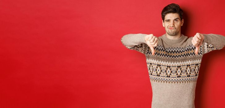 Portrait of skeptical and disappointed handsome man in christmas sweater, dislike new year party, showing thumbs-down, express disapproval, standing over red background.