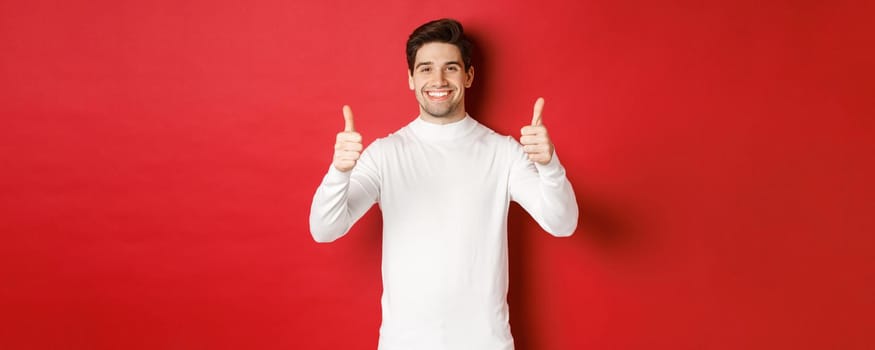 Concept of winter holidays, christmas and lifestyle. Portrait of satisfied handsome man in white sweater showing thumbs-up, praise something good, like it, standing over red background.