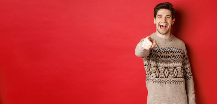 Concept of christmas celebration, winter and lifestyle. Happy attractive man in xmas sweater, wishing happy holidays and pointing at camera, standing over red background.