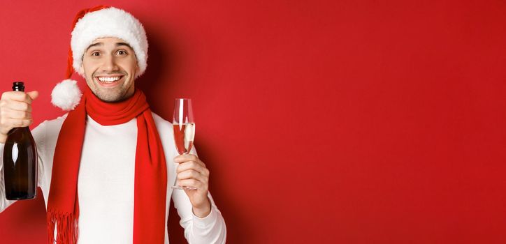 Concept of winter holidays, christmas and lifestyle. Close-up of cheerful handsome man, holding champagne bottle and glass, making toast for new year and celebrating, red background.