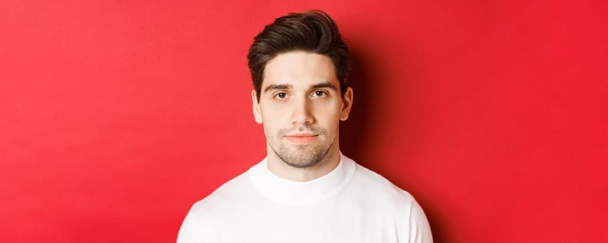 Close-up of handsome brunette guy in white sweater, smiling at camera, standing over red background.