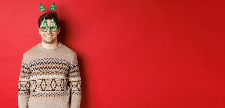 Portrait of happy and cute man in winter sweater and party glasses, celebrating new year or christmas, smiling while standing over red background.