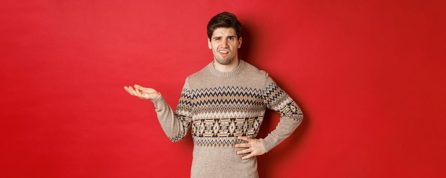 Image of confused and disappointed handsome man, complaining about christmas party, raising hand and looking bothered, standing over red background.