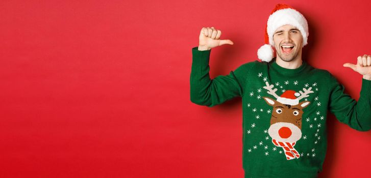 Concept of winter holidays, christmas and lifestyle. Sassy handsome man in santa hat and green sweater, pointing at himself and winking, standing over red background.
