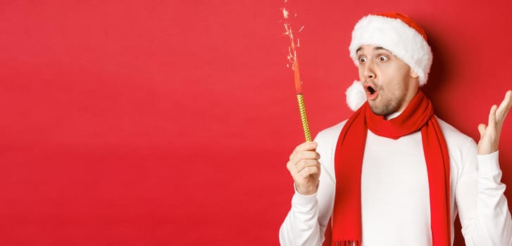 Concept of christmas, winter holidays and celebration. Close-up of amazed handsome man in santa hat and scarf, looking at sparkler with excitement, standing over red background.