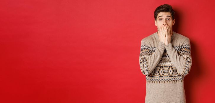 Portrait of shocked and speechless caucasian guy in christmas sweater, reacting to announcement, standing startled against red background.