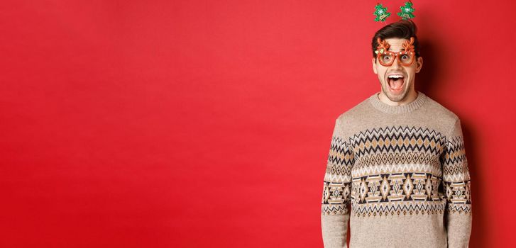 Image of surprised and excited handsome man, wearing party glasses with christmas sweater, drop jaw and looking at amazing new year promo offer, standing against red background.