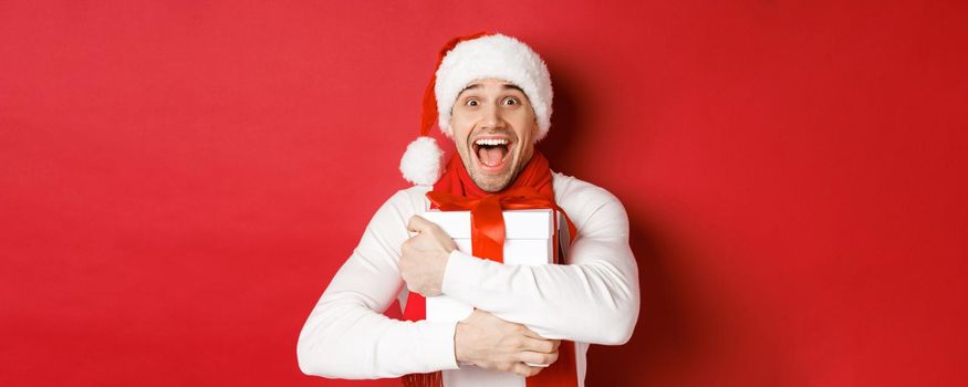 Concept of winter holidays, christmas and lifestyle. Close-up of happy man in santa hat receiving present, looking happy and hugging gift box, standing over red background.