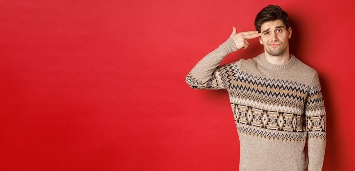 Portrait of distressed and annoyed handsome man, wearing christmas sweater, making finger gun sign near head and shooting himself, standing over red background.