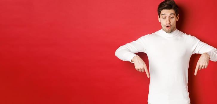 Portrait of surprised good-looking guy in white sweater, looking and pointing fingers down at logo, standing against red background.