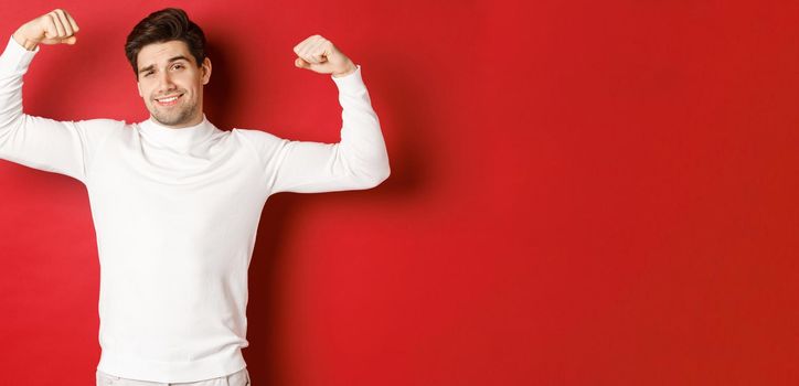 Portrait of smiling handsome man in white sweater, flexing biceps and bragging with strength, show-off strong muscles after workout, standing over red background.