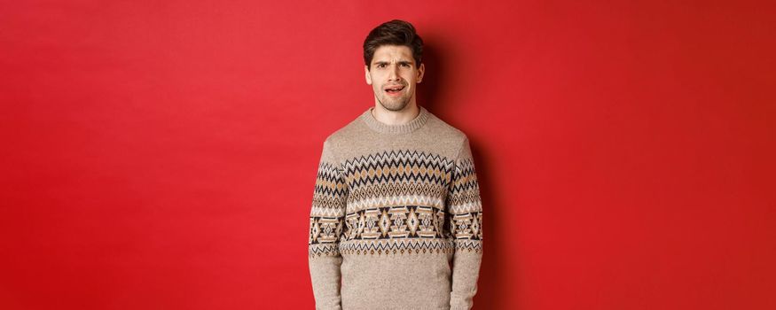 Portrait of confused adult man in christmas sweater, standing disappointed during new year holidays, dont understand something, standing over red background.