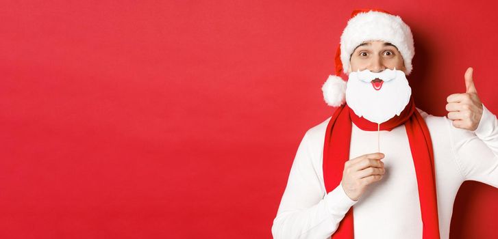 Concept of christmas, winter holidays and celebration. Portrait of funny man in santa hat, holding beard mask, showing thumb-up, enjoying new year party, standing over red background.