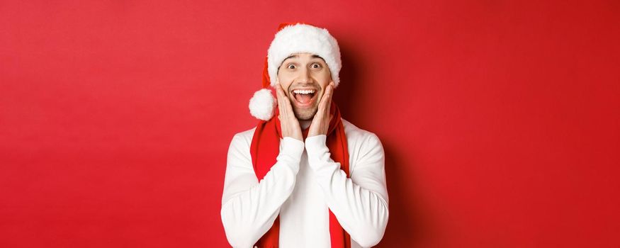 Concept of christmas, winter holidays and celebration. Close-up of surprised and happy man in santa hat and scarf, looking at something amazing, standing over red background.