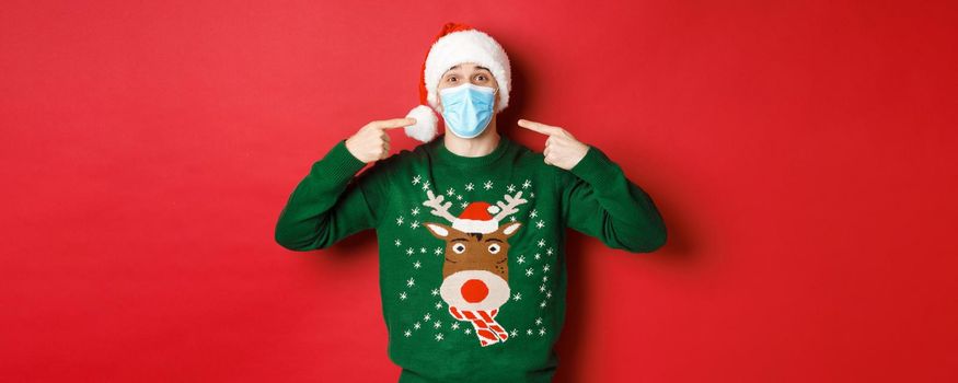 Concept of new year, coronavirus and social distancing. Happy man in santa hat and christmas sweater, recommending to wear medical mask on party, standing over red background.