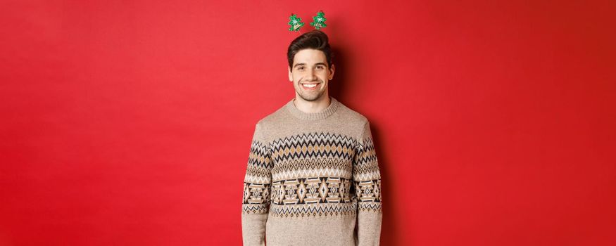 Concept of winter holidays, christmas and celebration. Handsome bearded guy in sweater wishing happy new year, smiling at camera, standing over red background.