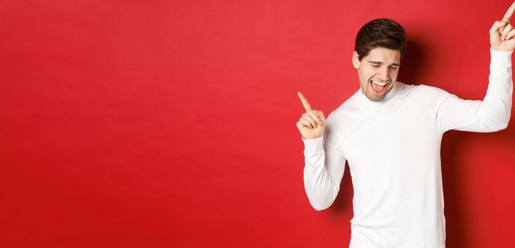 Portrait of handsome man enjoying new year party, dancing and having fun, pointing fingers up, standing over red background.